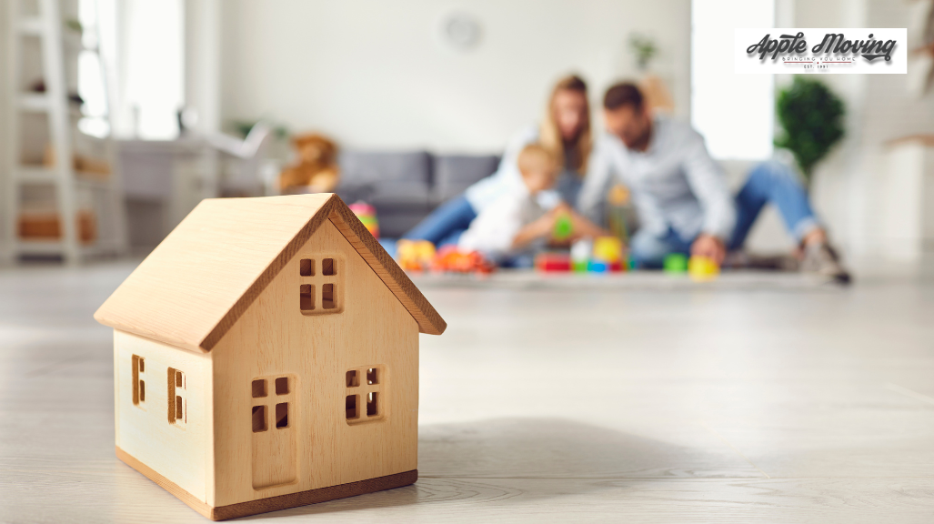 brown wooden miniature house in front of family of 3 sitting on the floor